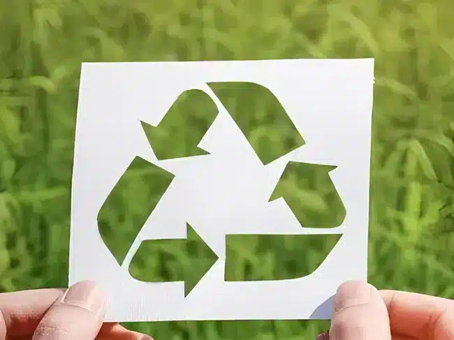A white square card with a cutout recycling symbol, held by two hands against a blurred green outdoor background.