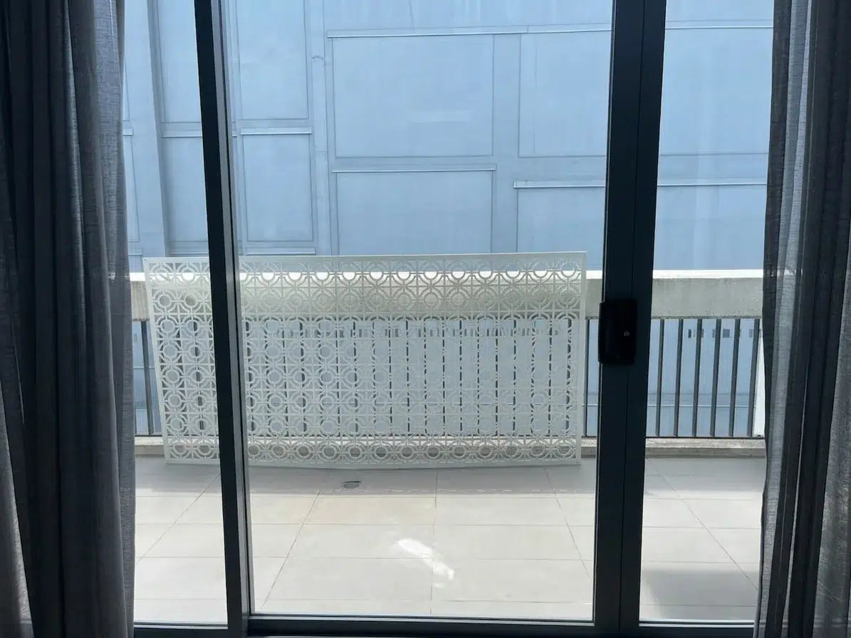 A white decorative outdoor railing panel with a geometric cutout pattern, installed on a balcony viewed through dark-framed sliding glass doors.