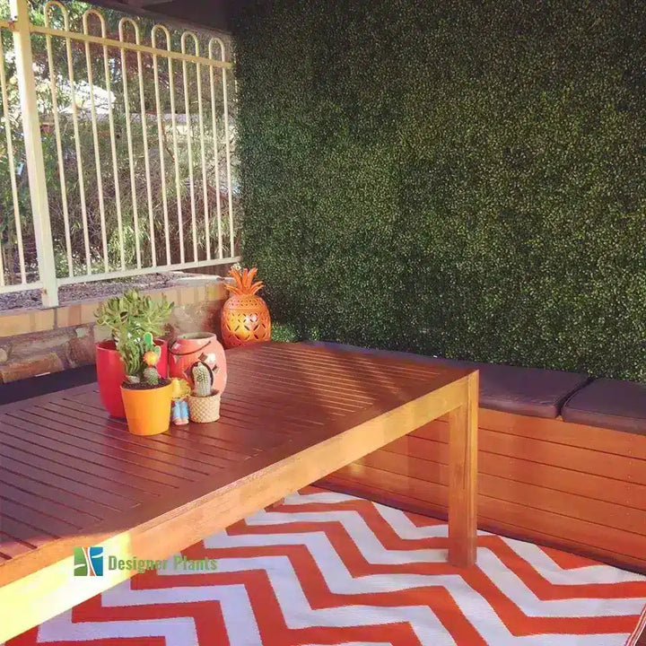 Outdoor patio furniture set featuring a rectangular wooden dining table, a built-in wooden bench with dark cushions, a green artificial hedge wall, and a red and white chevron patterned rug.
