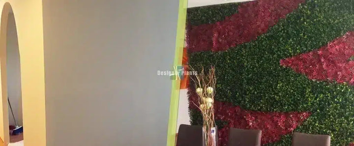 A decorative artificial green wall with bold red and green wavy patterns, shown in an interior setting with a vase of dried stems and chairs in front.