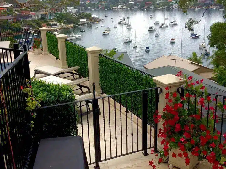 A scenic view from a modern patio overlooking a harbor filled with boats, with black metal railings, cream pillars, green hedges, lounge chairs, and red flowers in the foreground.