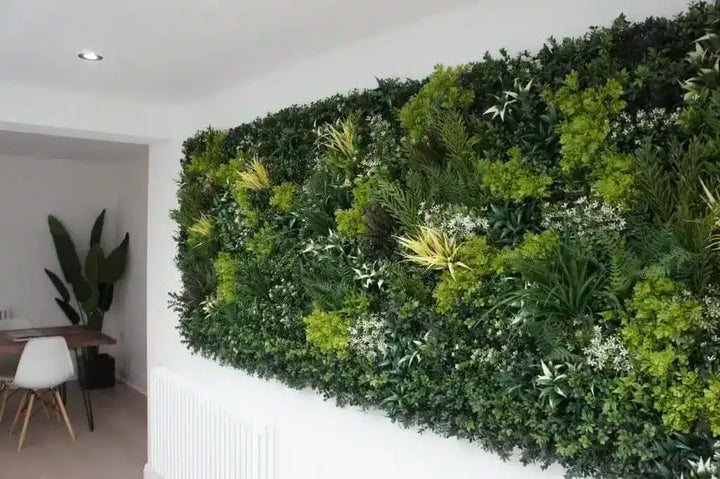 A large rectangular artificial green wall made of dense faux foliage and plants, mounted on a white wall in a modern interior room with a doorway and chair visible on the left.