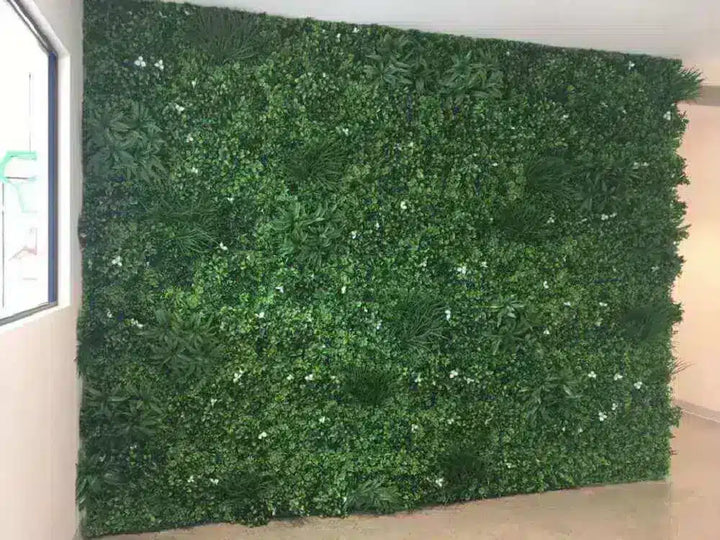 A large artificial green foliage wall panel with dense faux leaves and small white flower accents, shown installed against an indoor wall.