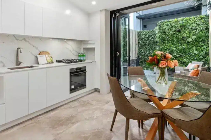 A modern kitchen and dining area with white cabinetry, a marble backsplash, a built-in oven, and a glass-top dining table with brown upholstered chairs. Large sliding glass doors open onto a patio with a green hedge background.