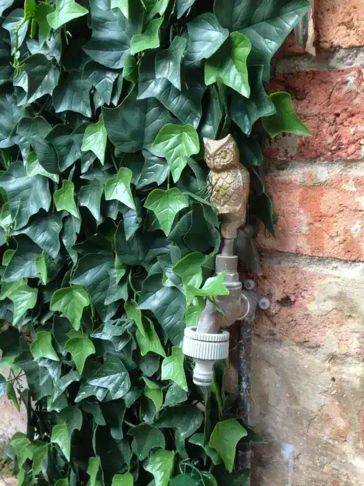 A close-up outdoor shot of a brass owl-shaped faucet handle on a metal spout, partially obscured by lush green ivy growing against a red brick wall.