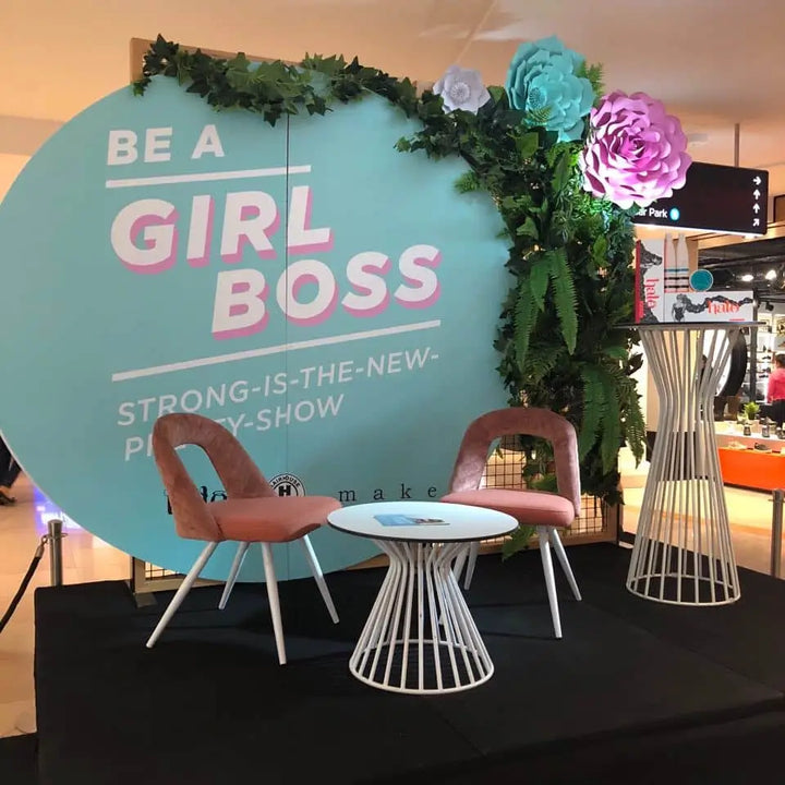 Decorative setup with 'Be a Girl Boss' sign, chairs, and floral arrangements in a store setting.