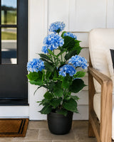 A potted artificial hydrangea with vibrant blue flowers and green leaves, displayed on a stone tile porch next to a wooden chair.
