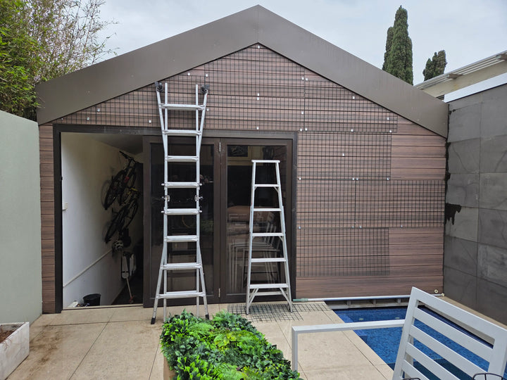 A modern outdoor shed or garage with brown wood-look cladding and a dark roof, shown with two silver extension ladders set up on a patio next to a swimming pool.