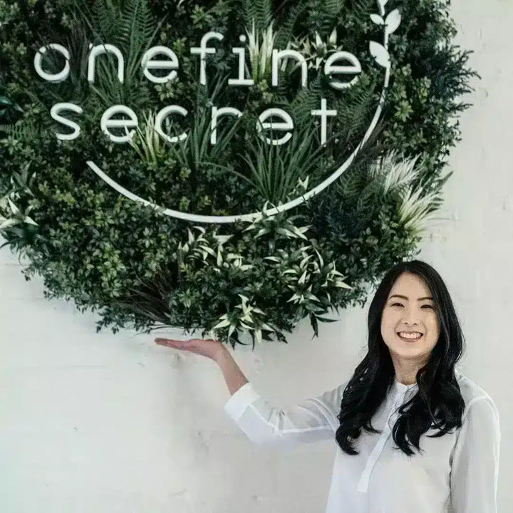 A smiling woman in a white blouse stands beside a large green leaf wall sign reading 'onefine secret' on a white wall.