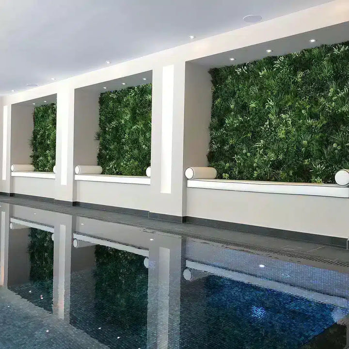 An indoor swimming pool with a modern design, featuring built-in white benches with cylindrical cushions and lush green vertical living walls behind them. The pool water is calm and reflects the ceiling lights and greenery.