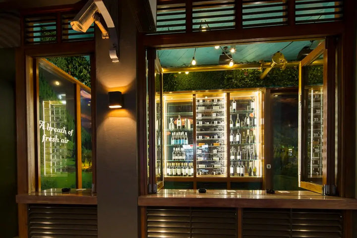 A warm, inviting bar or restaurant interior with a large glass window showcasing illuminated liquor cabinets filled with bottles. The scene features dark wood paneling, brass fixtures, and a greenery accent wall in the background, with a warm spotlight illuminating the bottles.