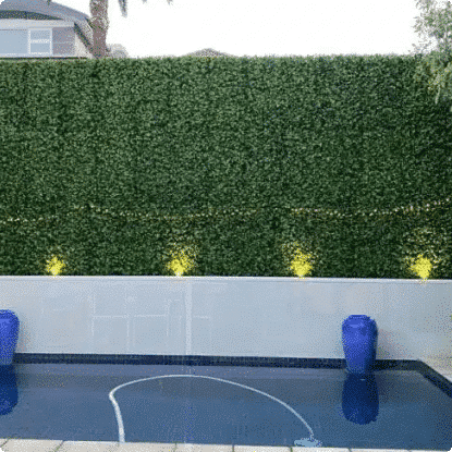 A tall, dense green artificial hedge wall installed behind a white retaining wall above a swimming pool, with blue decorative vases in the water and small yellow ground lights along the base.