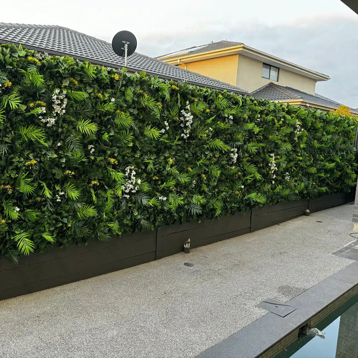 A long vertical green wall garden with dense artificial-looking palm leaves and scattered white and yellow flowers, installed along the side of a house above a paved walkway and small pool.