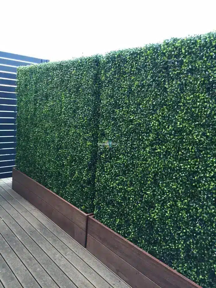 Green artificial hedge panel wall mounted on a dark brown wooden base, shown on an outdoor wooden deck next to a blue slatted fence.