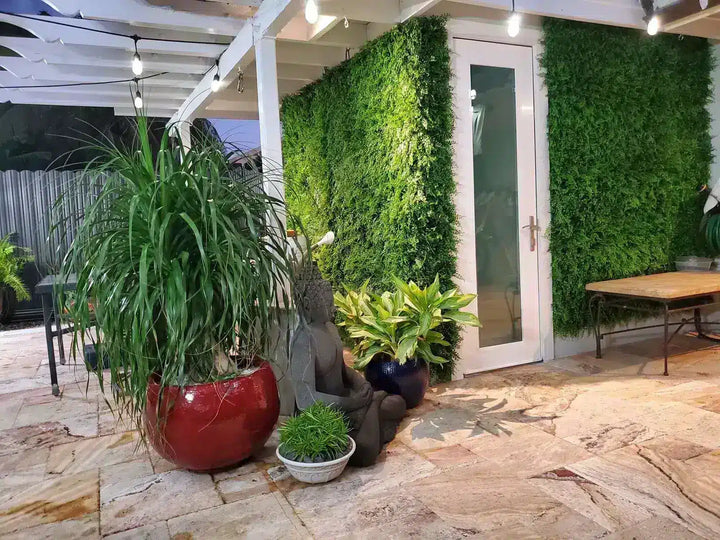 Outdoor patio scene with a large red planter holding tall green grass, a gray seated statue, smaller potted plants, a glass door, and artificial green wall panels under a covered patio.