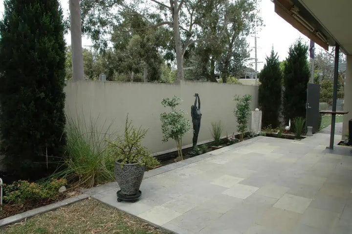A paved outdoor patio area with a light beige stucco wall, landscaped plants, a dark metal sculpture, and tall trees in the background.