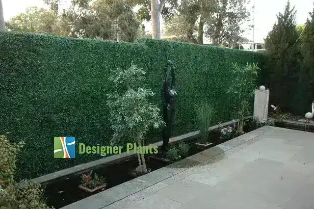 A large, dense green artificial hedge or privacy screen installed along the side of a paved patio, with small plants and a decorative black statue in a garden bed. A 'Designer Plants' logo watermark is visible in the lower-left corner.