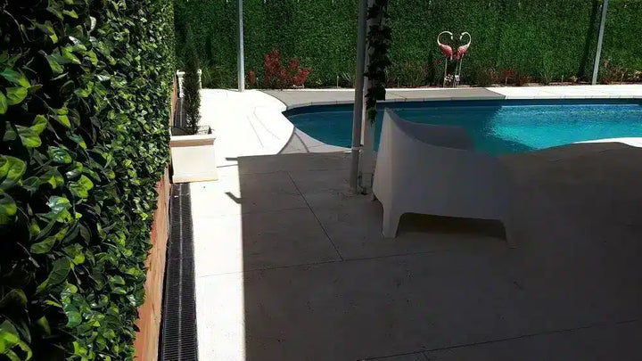 A modern outdoor swimming pool area with a curved white lounge chair in the foreground, a tiled deck, dense green hedges, and a flamingo-shaped pool float in the background.