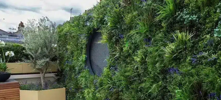 A lush vertical garden wall with dense green foliage, including palm-like plants and small purple flowers, covering a dark gray brick wall. A small tree, planter, and outdoor architectural elements are visible in the foreground and background.