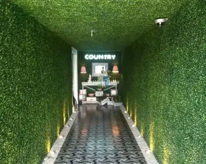 A narrow hallway with walls and ceiling covered in green artificial foliage, with a patterned dark floor. A lit sign at the far end reads COUNTRY above a small display area.