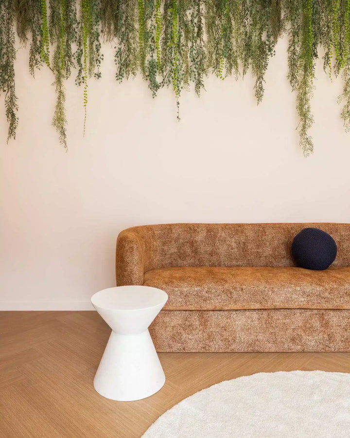 Brown sofa with a white side table and a black round pillow, against a light pink wall with green plants.
