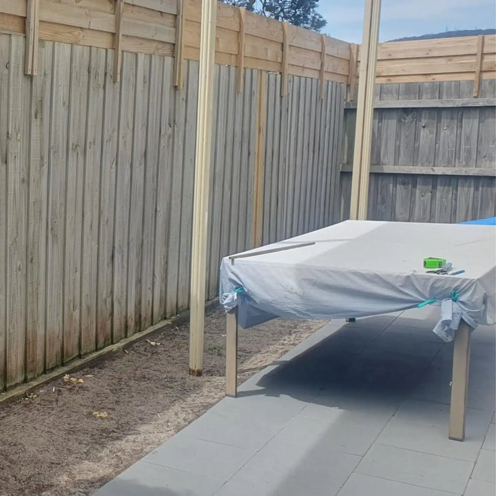 A covered table or bed with a light gray sheet and wooden frame, set outdoors on a paved patio beside a tall wooden fence.