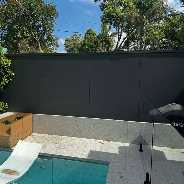 A modern outdoor backyard scene with a dark charcoal gray wall, a tiled pool deck, turquoise swimming pool water, a white slide, green trees, and blue sky.