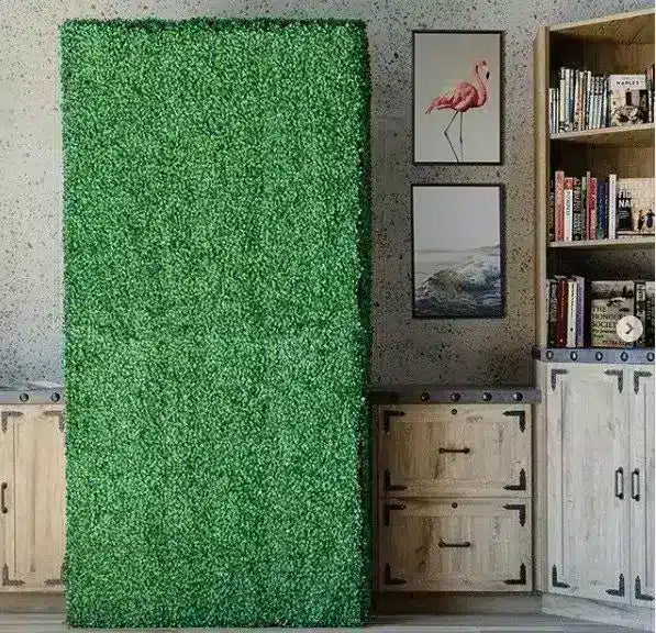 A tall rectangular green artificial grass or moss wall panel standing upright in an interior room next to a bookshelf, cabinet, and framed wall art.