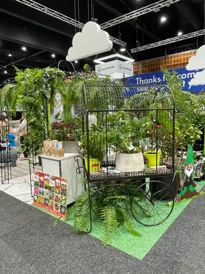 A decorative black metal cart styled with potted green plants, ferns, and artificial foliage, set up at an indoor exhibition or trade show with booths, signage, and ceiling truss structures visible.