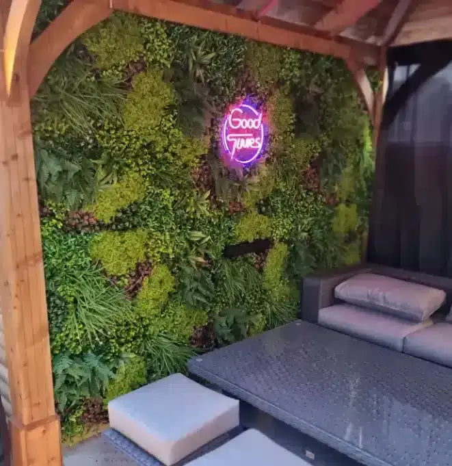 A lush green living wall with a glowing purple neon sign that reads 'Good Times' in a wooden covered patio setting with a dark gray outdoor sofa and cushions.