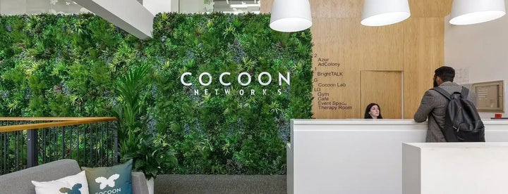 A modern office lobby with a large green living wall featuring the text 'COCOO NETWORKS' in white. A reception desk with a person working behind it and another person with a backpack standing at the counter are visible.