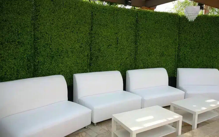A row of modern white outdoor lounge chairs and matching white coffee tables positioned in front of a tall green artificial hedge wall.