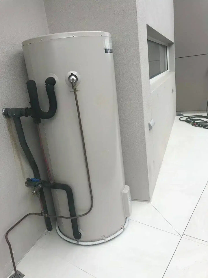A large cylindrical water heater or storage tank in a light gray/off-white finish, installed in a corner of a room with white tiled flooring and gray walls. Black plumbing pipes and a metal supply tube are attached to the side of the tank.