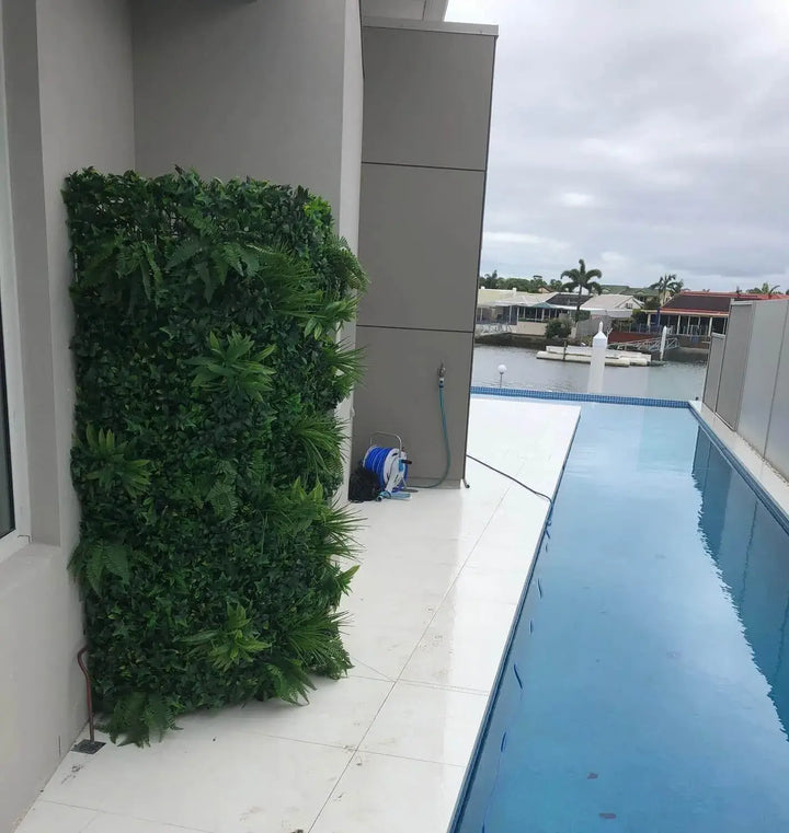 A tall rectangular artificial green wall hedge mounted on a white exterior wall next to a swimming pool, with a pool spigot and hose visible on the deck.