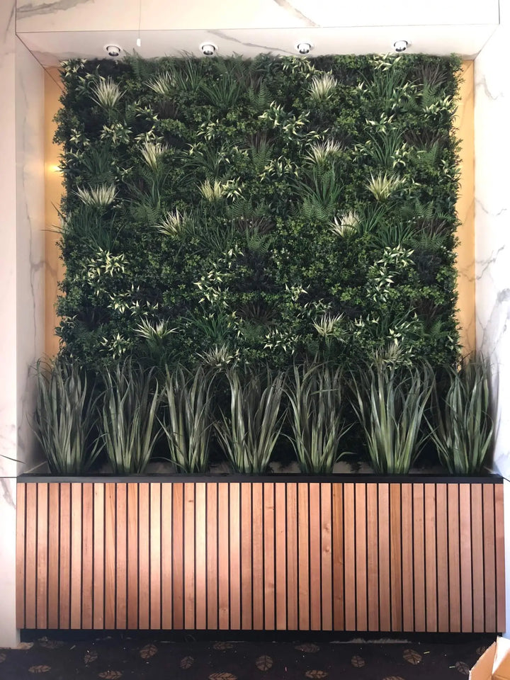A large artificial vertical green wall garden with dense green foliage and spiky grass-like plants, mounted above a slatted wood planter box with tall ornamental grasses in front.