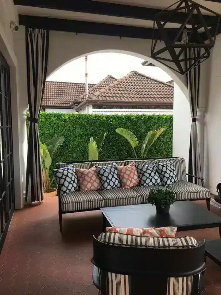 A modern outdoor patio setup featuring a striped sofa with patterned throw pillows, a matching armchair, a black coffee table, and a large arched opening looking out to a green hedge.