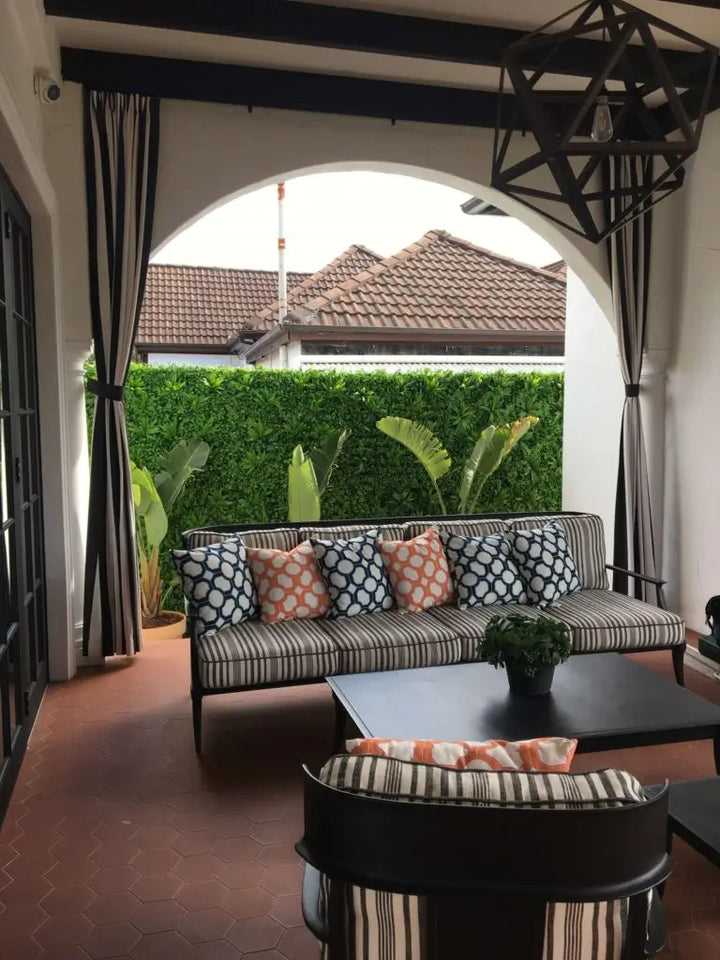 A modern outdoor patio seating area featuring a striped sofa with patterned throw pillows, a black coffee table, and matching striped chairs under a black geometric hanging light, set against a lush green hedge and tiled roof in the background.