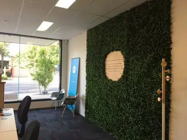 An office interior featuring a prominent artificial green hedge wall with a circular wooden plaque in the center. The room includes a large window looking out onto trees, dark carpet, office chairs, and a wooden floor rod.