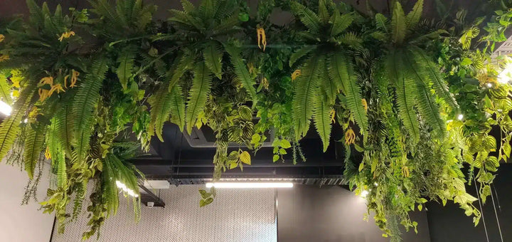A large hanging greenery wall feature with dense artificial ferns and trailing foliage suspended above a modern interior ceiling and wall.