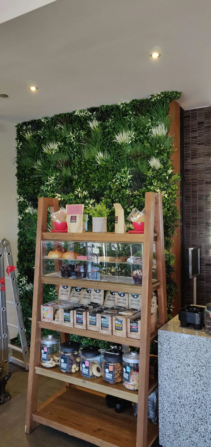 A wooden four-tier display shelf filled with packaged snacks, jars, and small potted plants, set in front of a green foliage wall in an indoor retail or cafe environment.