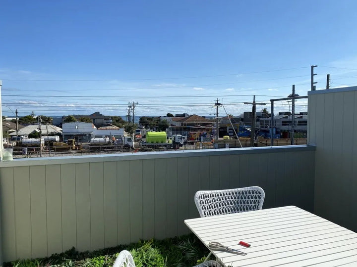 Outdoor balcony view with a white slatted table and chair in the foreground, overlooking a residential area with houses, utility poles, and the ocean under a blue sky.