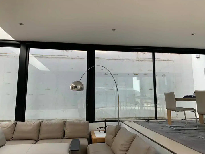 A modern living room interior featuring a light beige sectional sofa, a tall silver floor lamp, and large black-framed windows looking out onto a snowy outdoor wall.