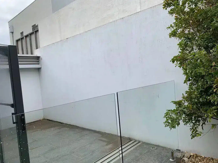 A modern outdoor patio area featuring a white stucco wall, a glass panel fence, gray stone flooring, and a green tree on the right.