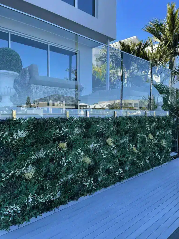 A modern glass fence with gold posts and a lush green living wall hedge on a blue wooden deck in front of a contemporary glass house.