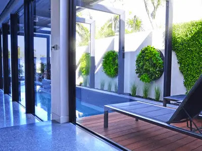 Modern outdoor patio area with a wooden deck, glass sliding doors, a swimming pool, and a lounge chair beside green wall planters and hedges.