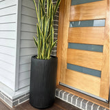 Tall black ribbed planter with snake plant by a modern wooden front door.