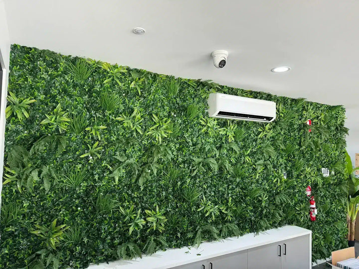 A large artificial green foliage wall mounted on an office wall, featuring various shades of green plants and leaves. A white air conditioner is mounted on the wall above a white cabinet, and a security camera is visible on the ceiling.