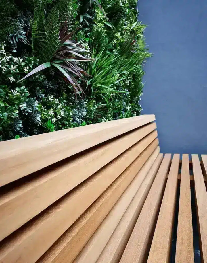 A close-up view of a modern wooden slat bench or deck seat with a green living wall and a dark blue-gray background.