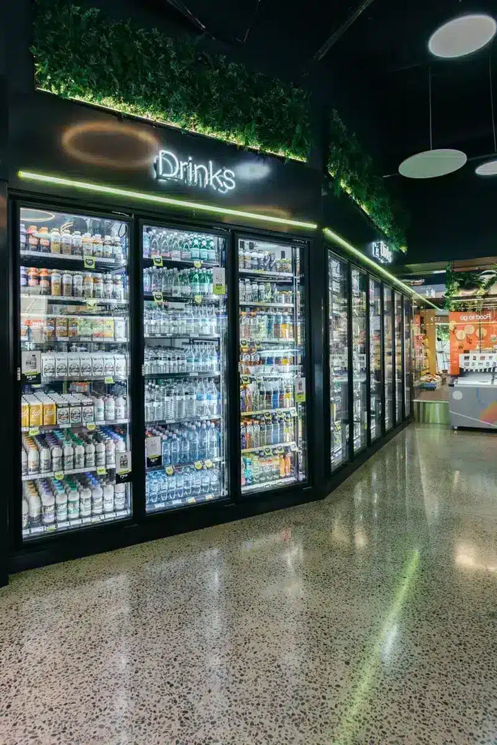 A row of commercial glass-door beverage fridges stocked with bottled and canned drinks in a retail store. The units have black frames and doors, illuminated green LED lighting along the top, and a white 'Drinks' sign above the first unit.