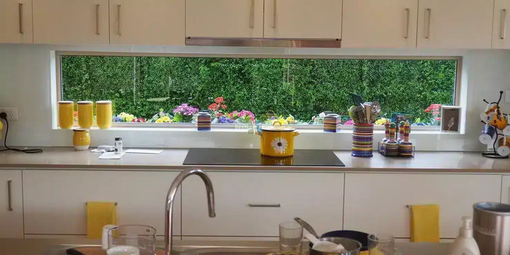 A modern kitchen interior with white cabinets, a stainless steel sink faucet, a black cooktop, and a large window looking out onto a green hedge wall with colorful flowers.